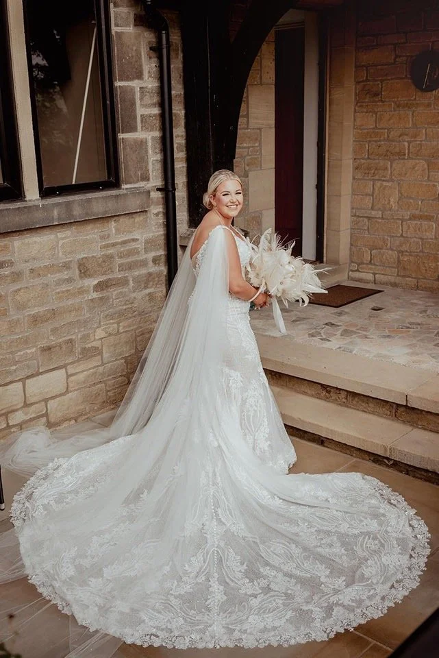 A bride in a white wedding gown holding a bouquet of feathers, standing outdoors on stone steps, smiling.