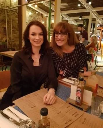 Two women sitting at a restaurant table, smiling at the camera. The table has condiments and menu items, and there are other people dining in the background.