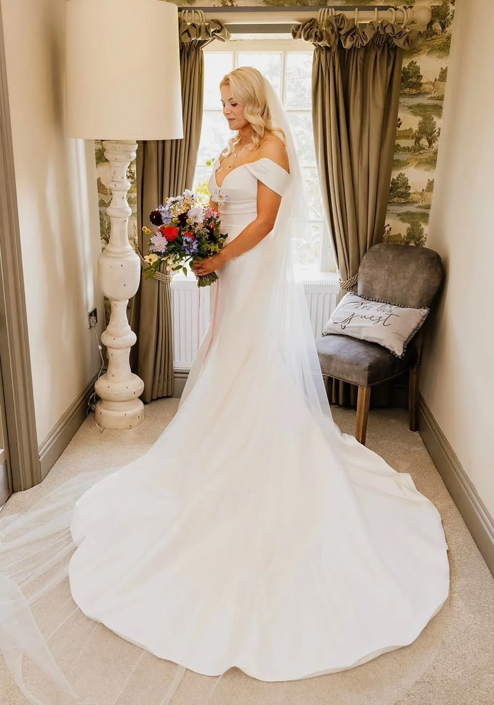 A bride in a white wedding gown holding a colorful bouquet of flowers standing in a cozy, well-lit room with beige walls, a window with curtains, a gray upholstered chair with a pillow, and a tall white floor lamp.