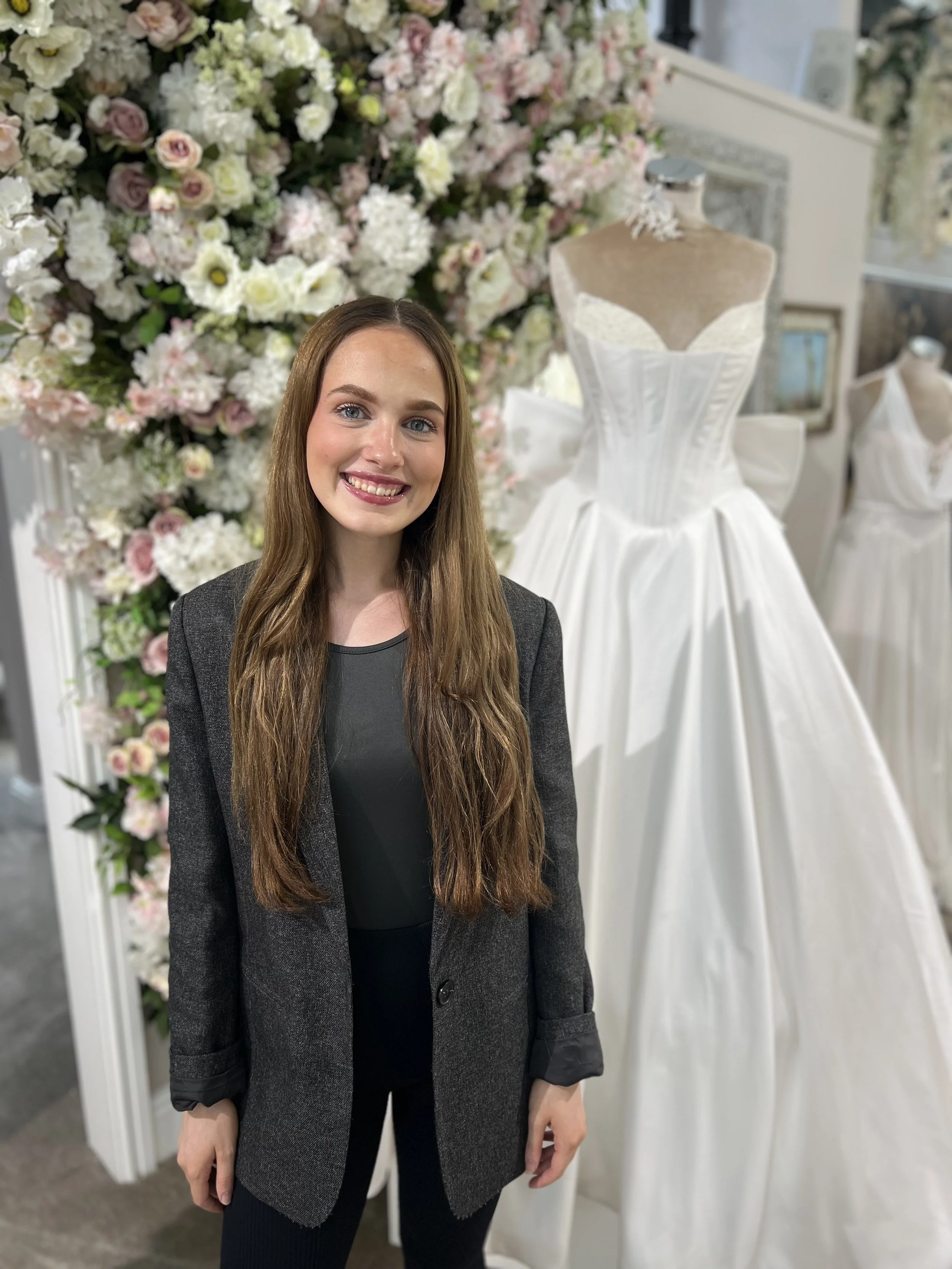 A woman with long brown hair smiling in front of wedding dresses and a floral arrangement.