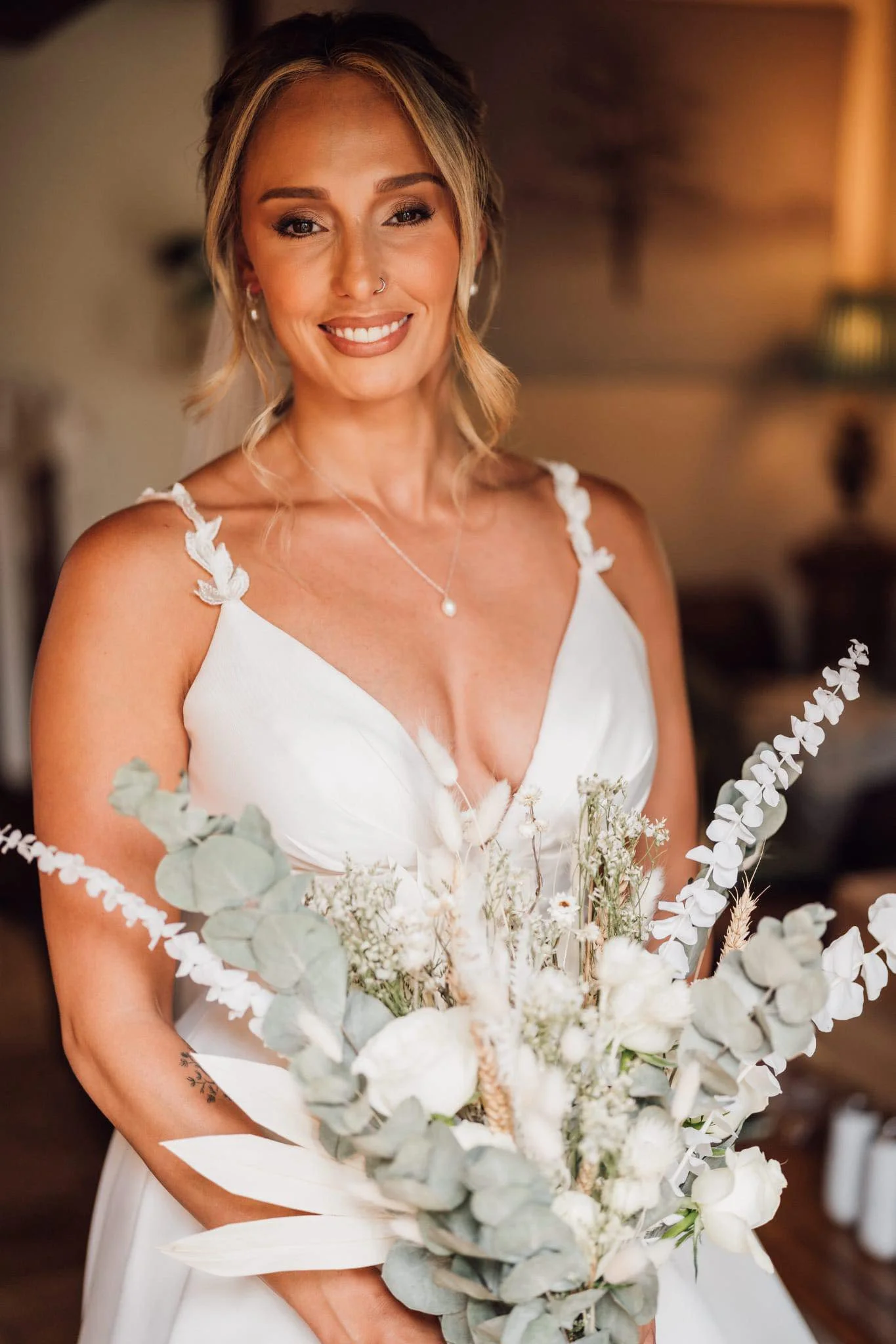 A smiling woman in a white dress holding a bouquet of white and green flowers, with shoulder-length blonde hair, a nose ring, and jewelry, indoors with warm lighting.