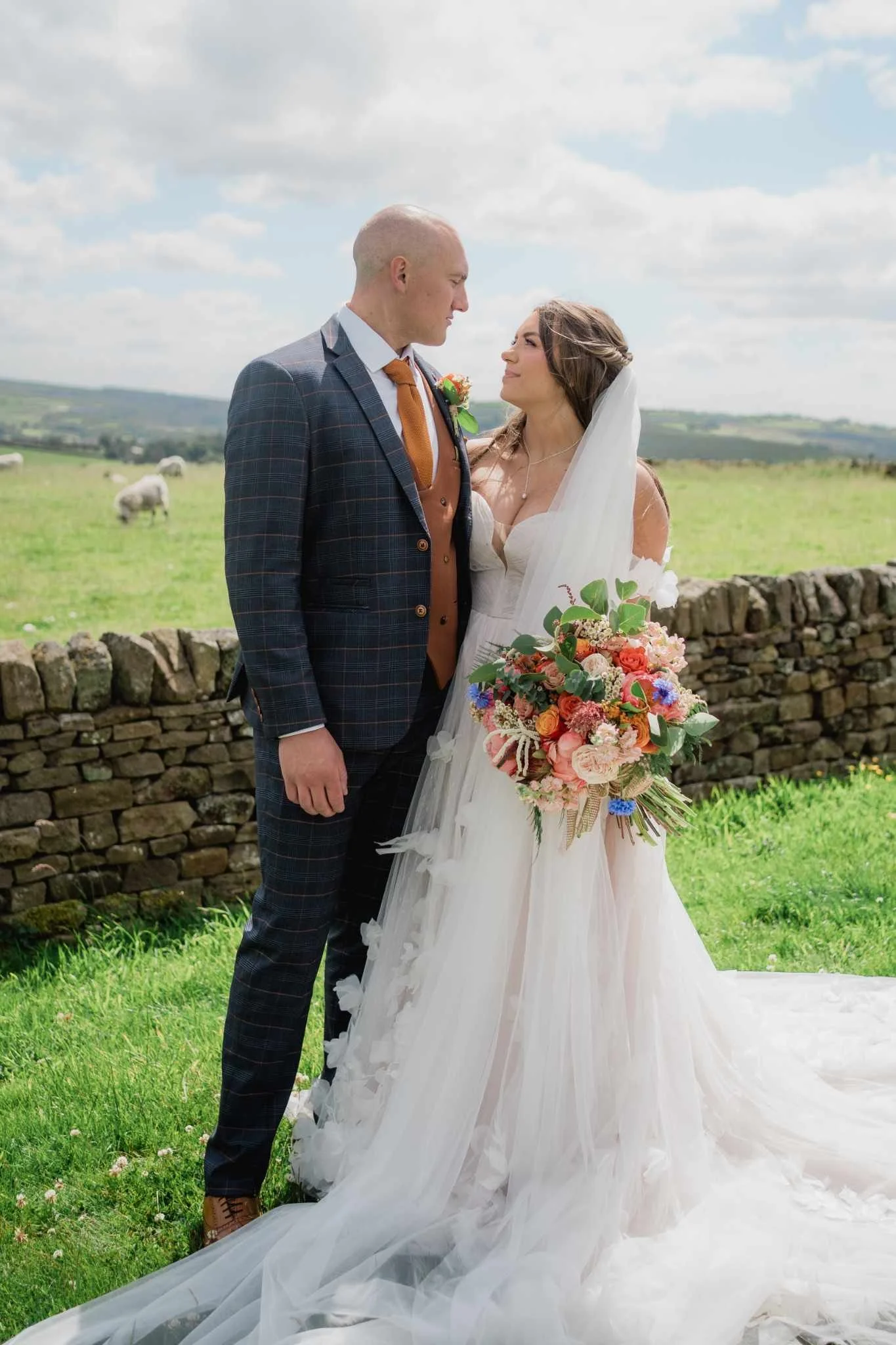 A bride and groom stand outdoors in front of a stone wall, gazing into each other's eyes, with a lush green landscape and sheep in the background. The bride wears a white wedding gown and veil, holding a colorful bouquet. The groom wears a plaid suit