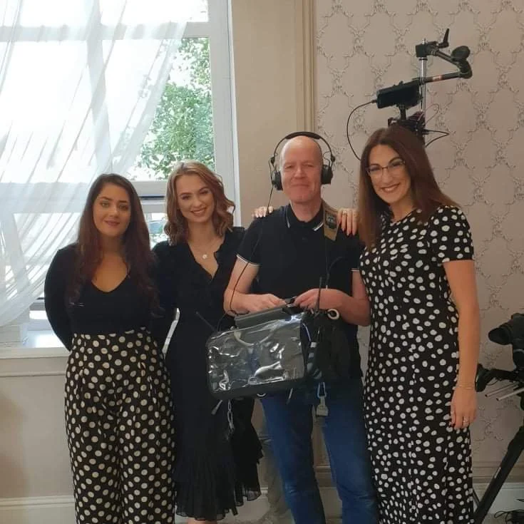 A group of four people, three women and one man, posing together indoors. The man is wearing headphones and holding professional camera equipment. The women are dressed in polka dot outfits, standing next to him.