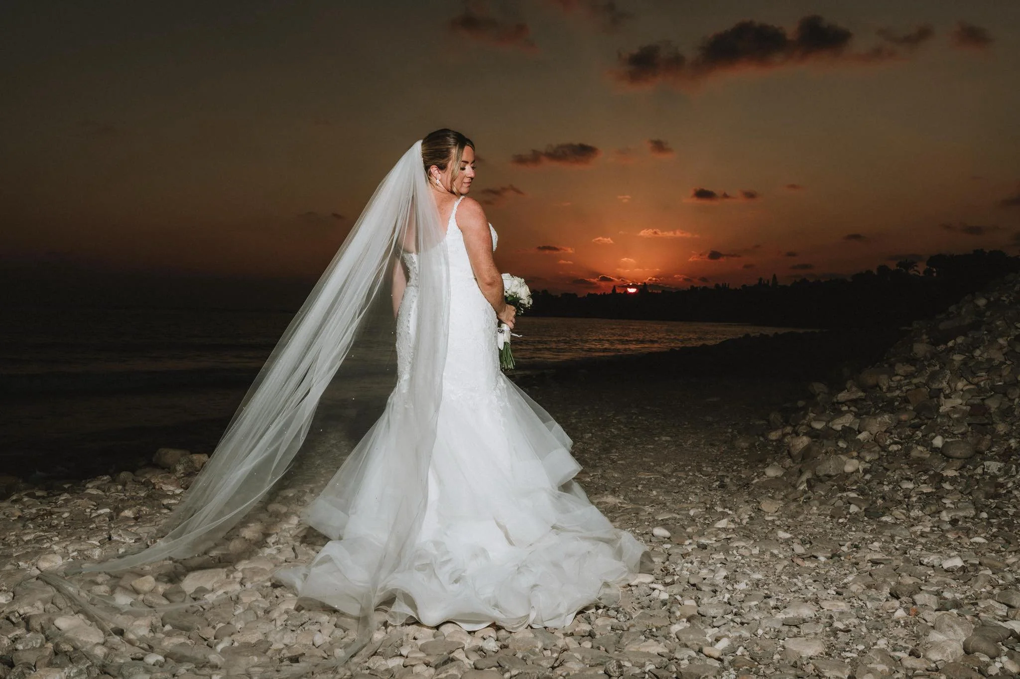 Bride in a white wedding gown and veil holding a bouquet, standing on rocky beach at sunset.