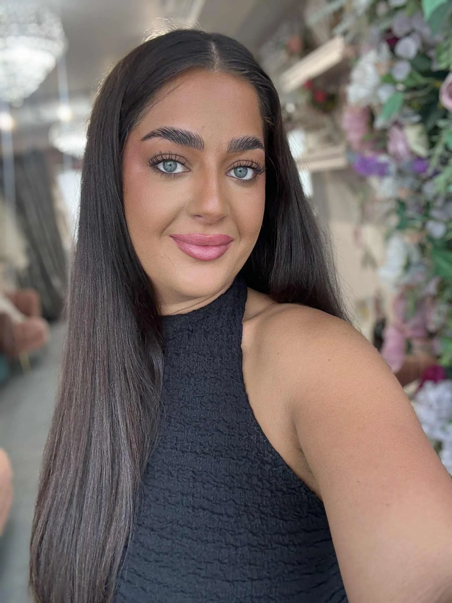 A woman with long dark hair, blue eyes, and light makeup, wearing a black sleeveless top, standing in front of a floral background and taking a selfie.