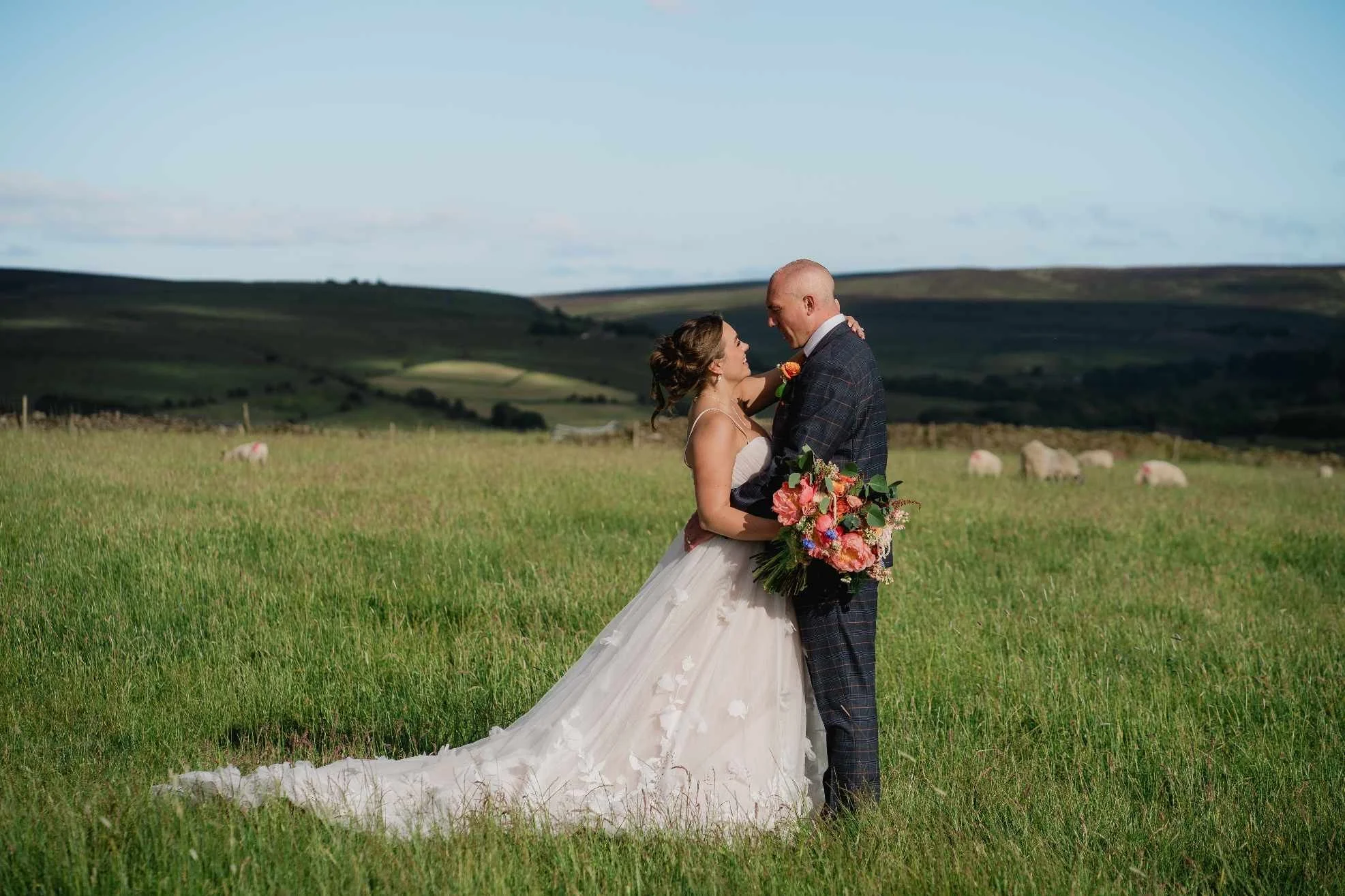 A bride and groom embrace in a green field with rolling hills in the background, with a clear blue sky above. The bride is in a white wedding gown holding a large bouquet, and the groom is in a dark plaid suit.
