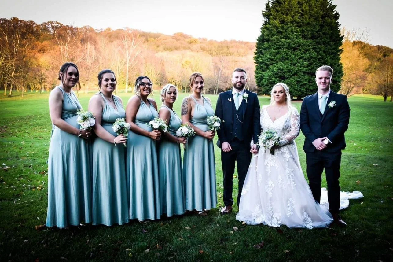 A wedding party standing in a grassy field during autumn, featuring six women in matching light blue dresses, a man and woman in wedding attire, and another man in a dark suit, all holding bouquets and posing for a photo.