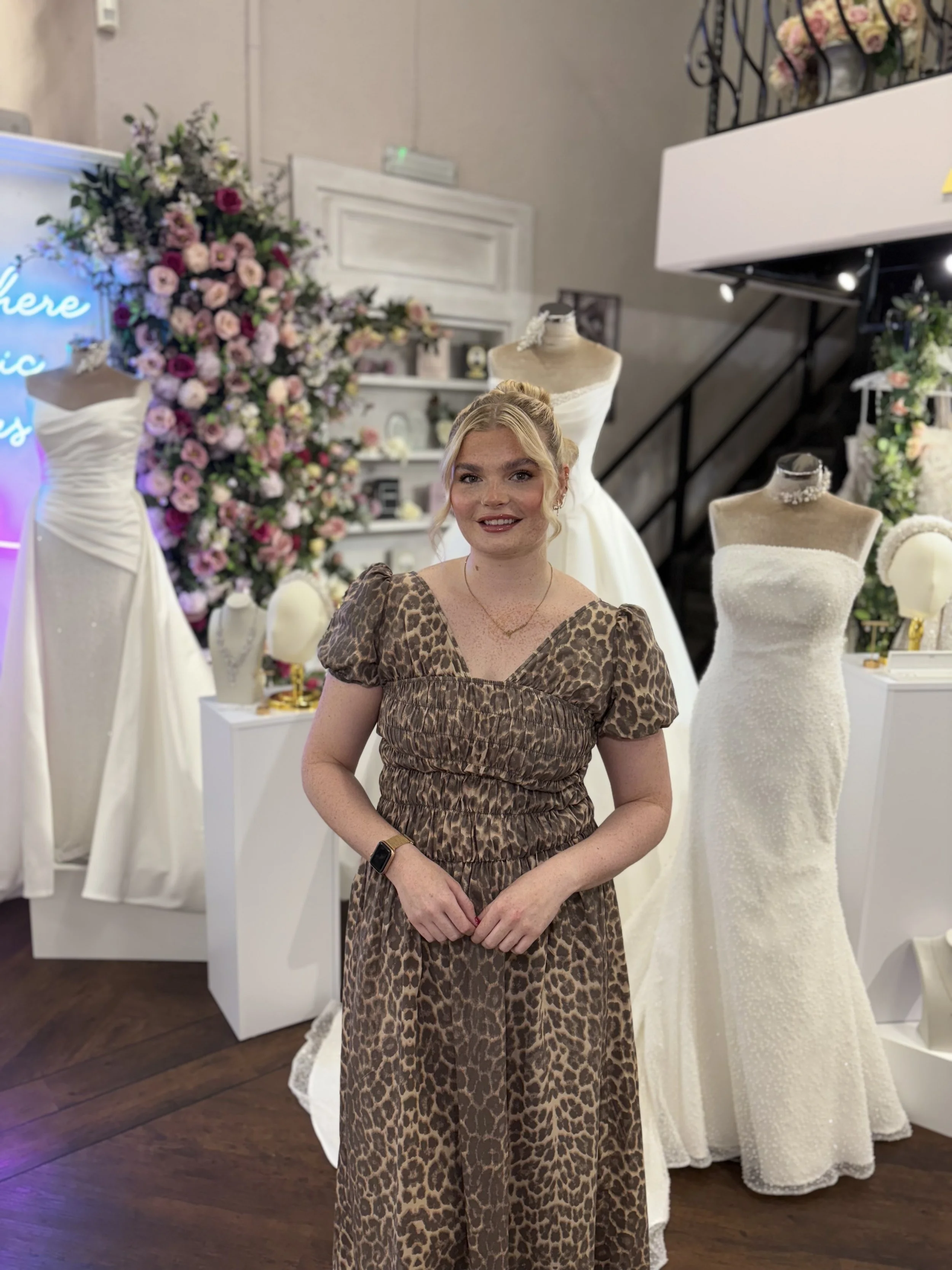 A woman standing in front of wedding dresses and mannequins in a bridal boutique, with a large flower arrangement in the background.
