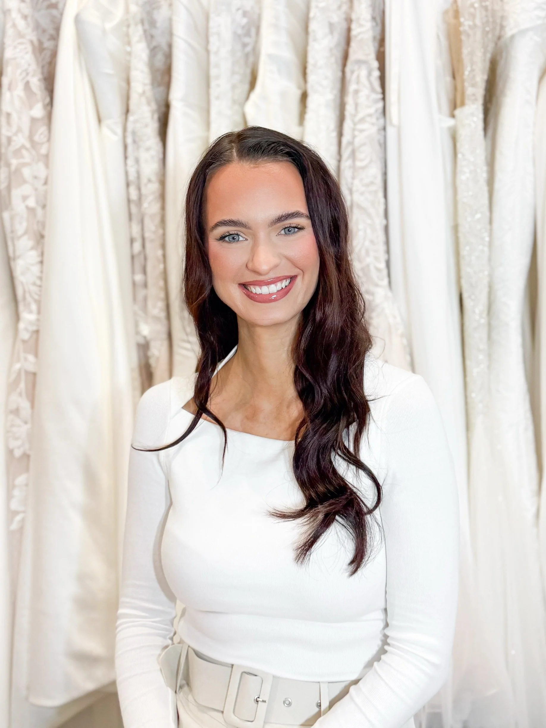 A woman with long dark hair and a black sleeveless top standing in front of a display of white wedding dresses and a bouquet of white and pink flowers on a marble table.