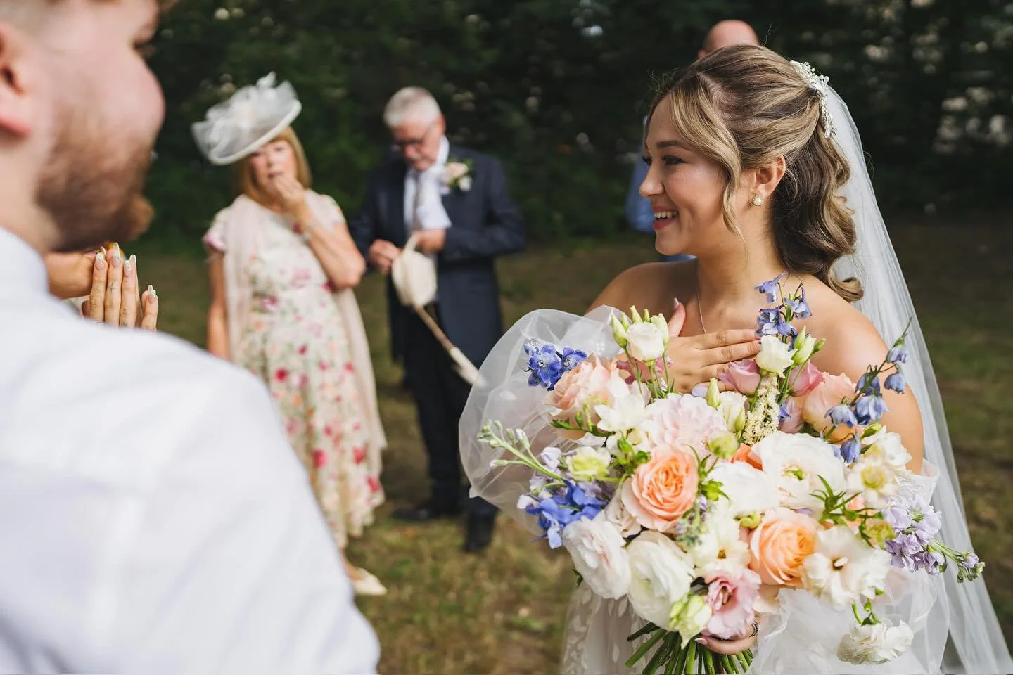 A bride dressed in a wedding gown holding a large bouquet of pink, white, and purple flowers, smiling and touching her chest while talking to a man in a white shirt during an outdoor wedding ceremony.