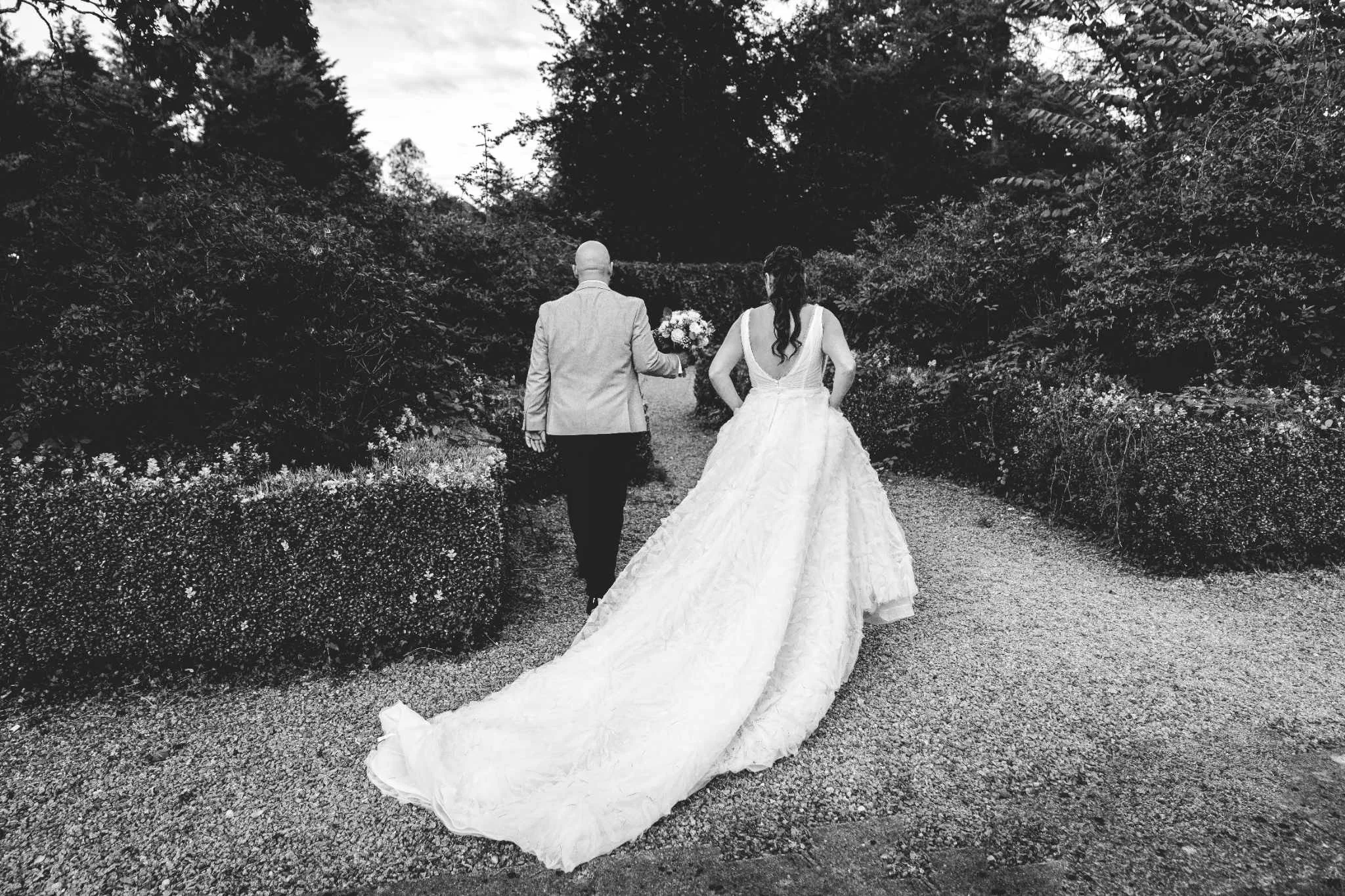Black and white photo of a bride in a long wedding dress and a groom in a suit walking down a garden path, holding a bouquet of flowers, with trees and bushes on either side.