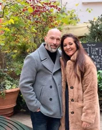 A man and woman smiling outdoors in a garden setting, with greenery and potted plants in the background.