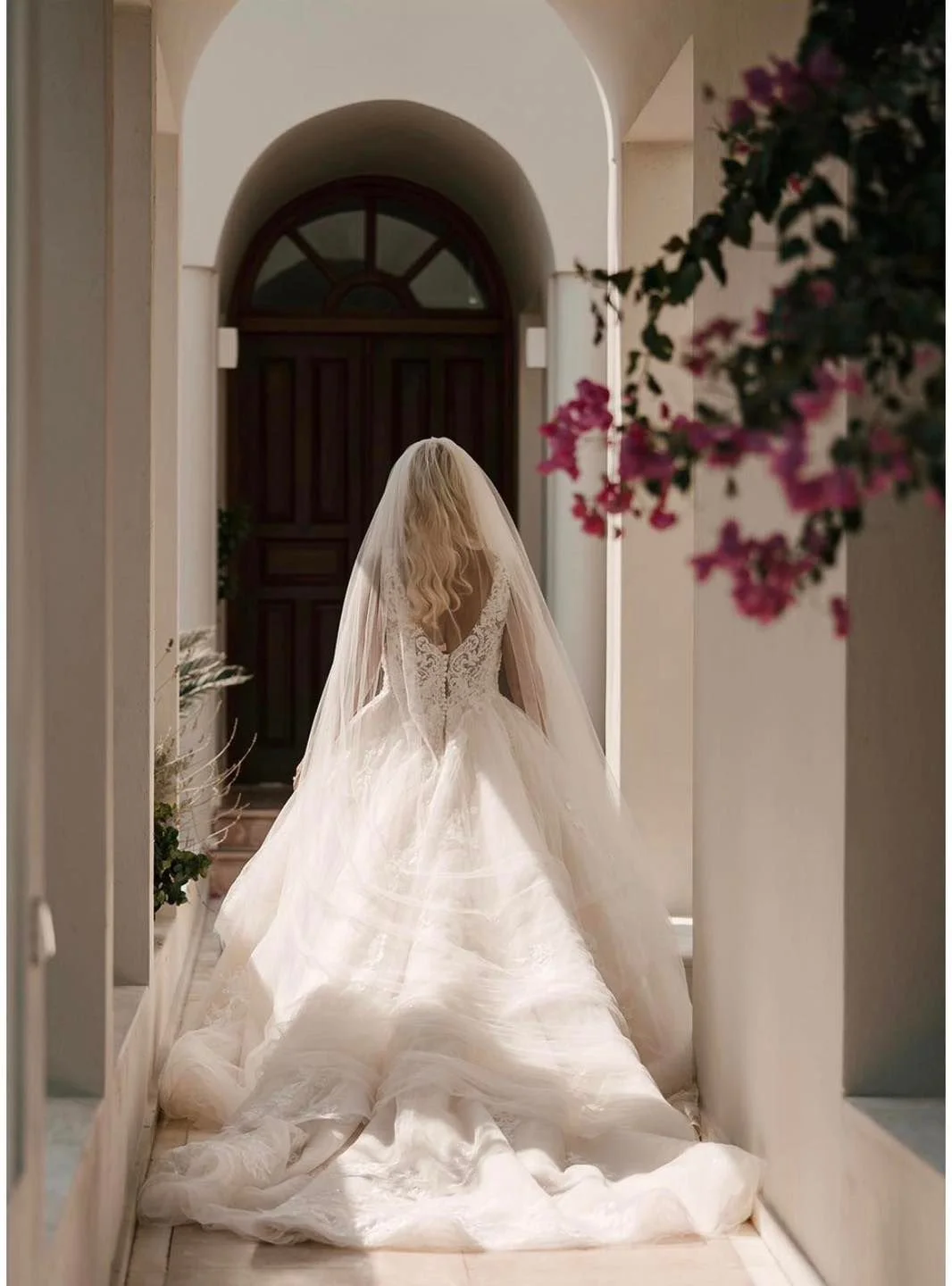 A bride in a white wedding gown with a long train and lace back, standing in a hallway with an arched door at the end, surrounded by pink flowers.