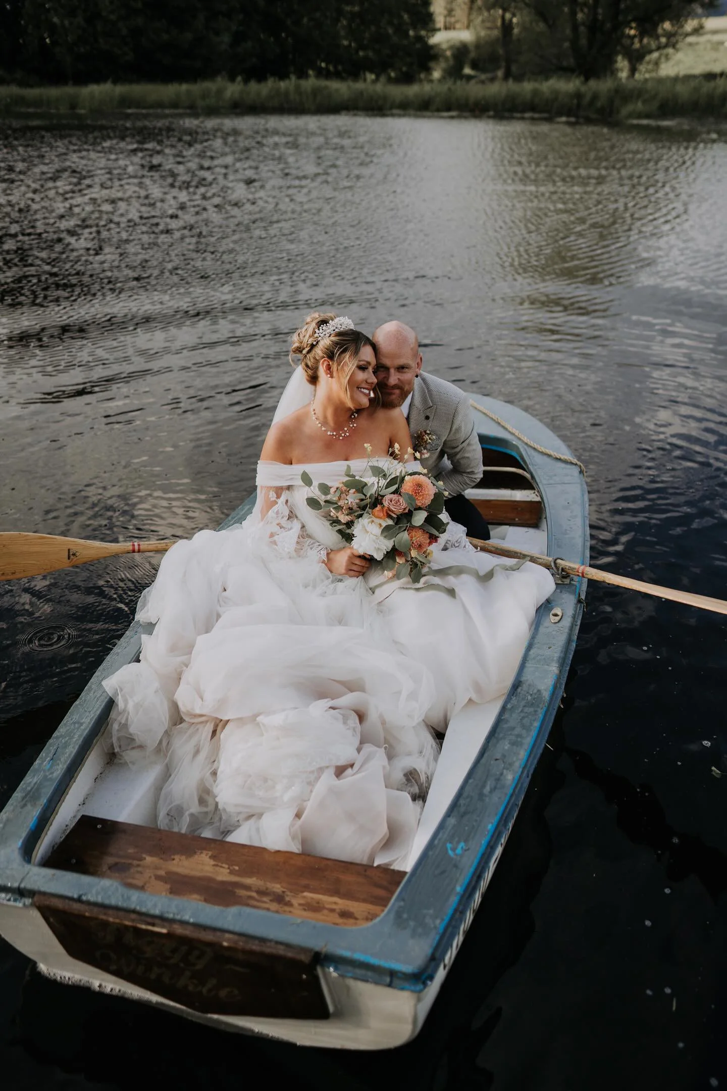 A bride and groom sitting in a boat on a lake during their wedding, with the bride holding a bouquet of flowers.