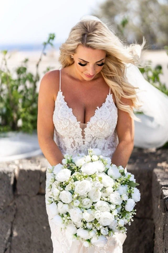 A bride with long blonde hair in a white wedding dress holding a large bouquet of white roses, looking down outdoors.