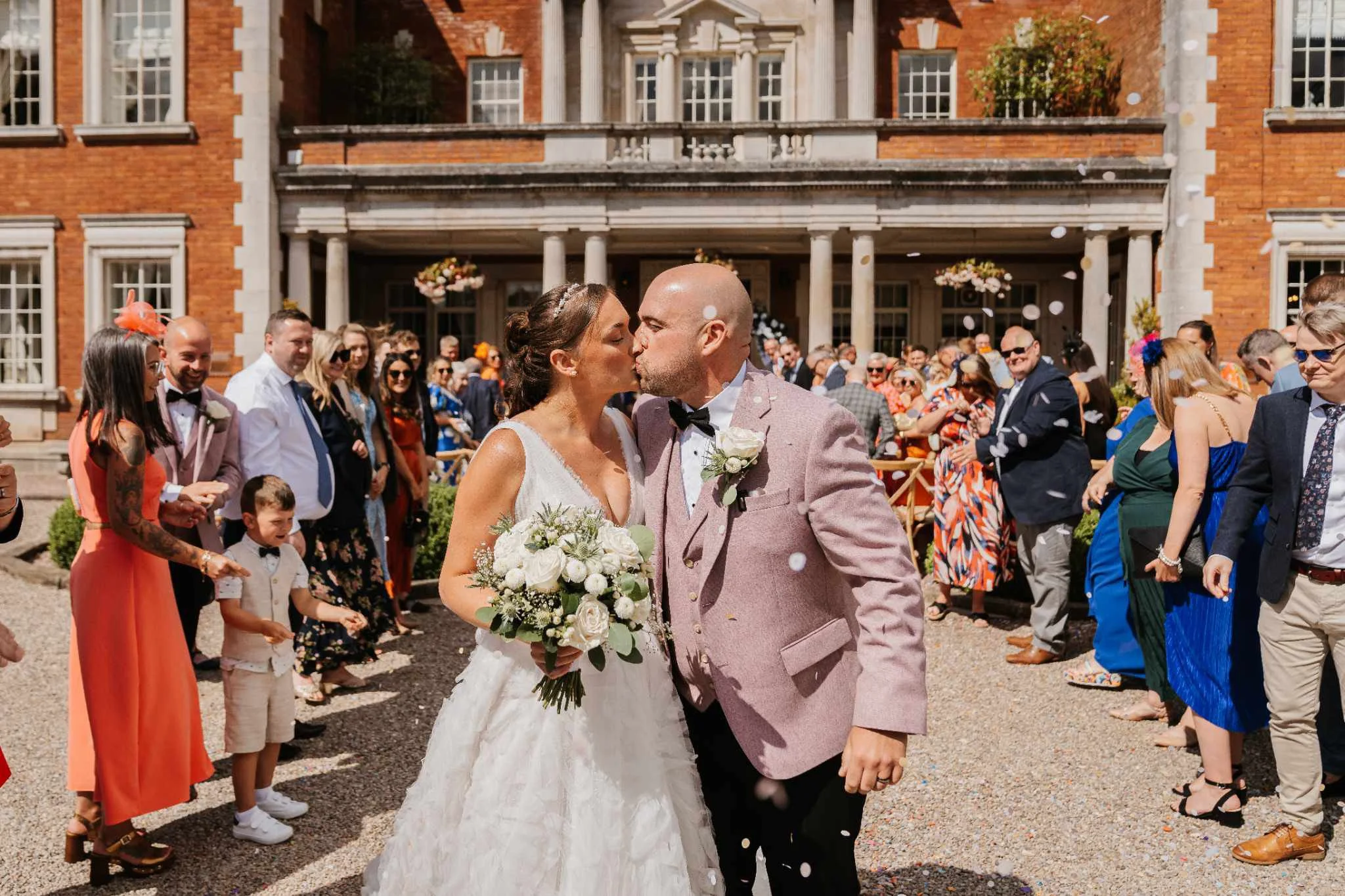 Bride and groom kissing outside a brick building with guests celebrating around them.