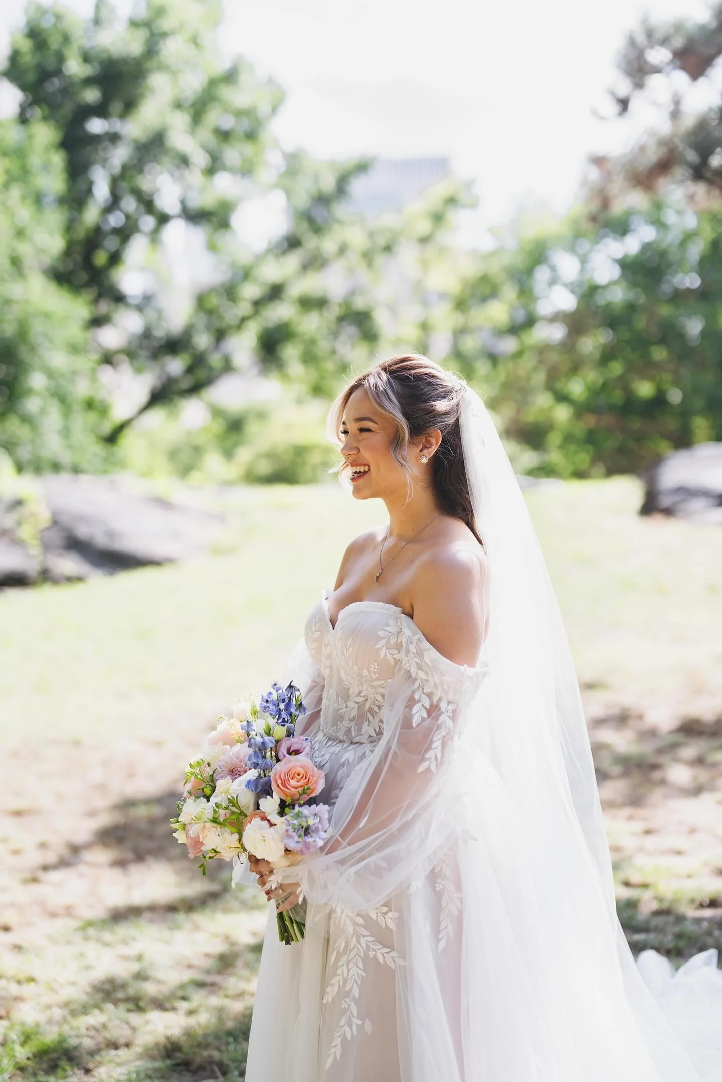 A smiling bride in a white wedding dress with floral embroidery, holding a bouquet of pink, purple, and white flowers, standing outdoors with green trees and sunlight.