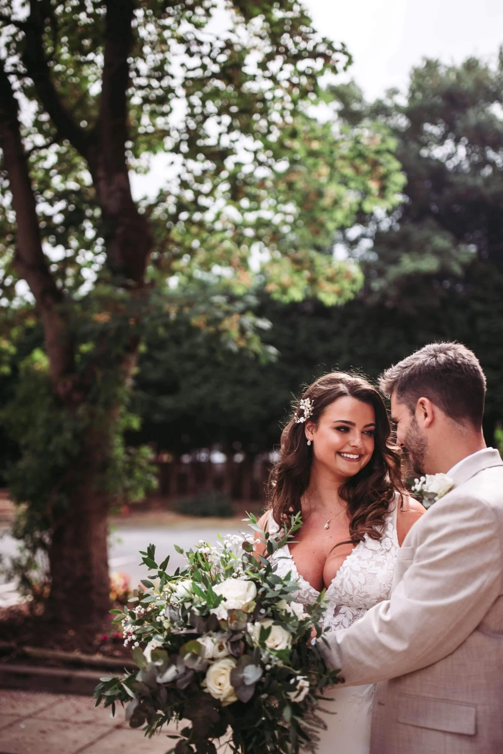 A bride and groom are looking at each other and holding a bouquet of white roses and greenery outdoors during daytime.