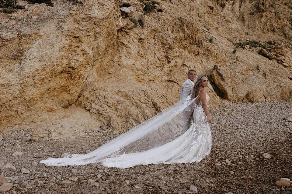 A bride and groom dressed in wedding attire standing on rocky terrain against a cliffside. The bride is wearing a white gown with a long veil, and the groom is in a light-colored suit. They are facing away from the camera, looking back over their sho