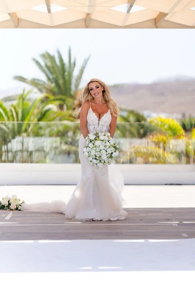 A bride in a white lace wedding gown holding a large bouquet of white flowers, standing outdoors with a palm tree and hilly landscape in the background.