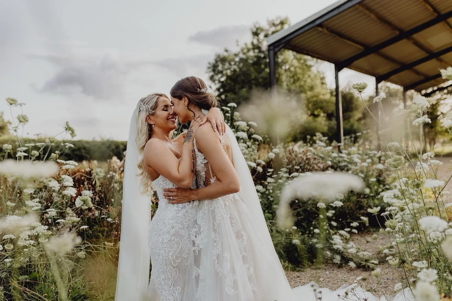 Two women in wedding dresses embracing in a flower garden, smiling and touching foreheads, with a pavilion and trees in the background.