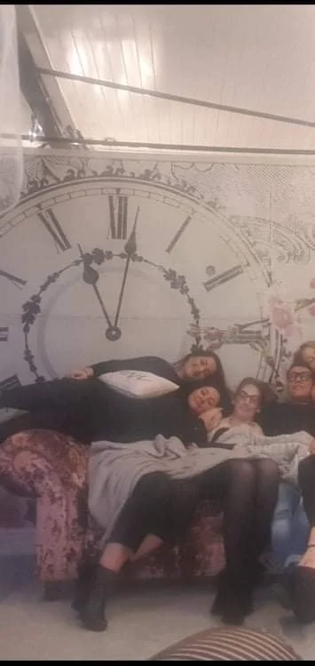 Four people sitting and lying on a vintage sofa in front of a large clock mural on the wall.