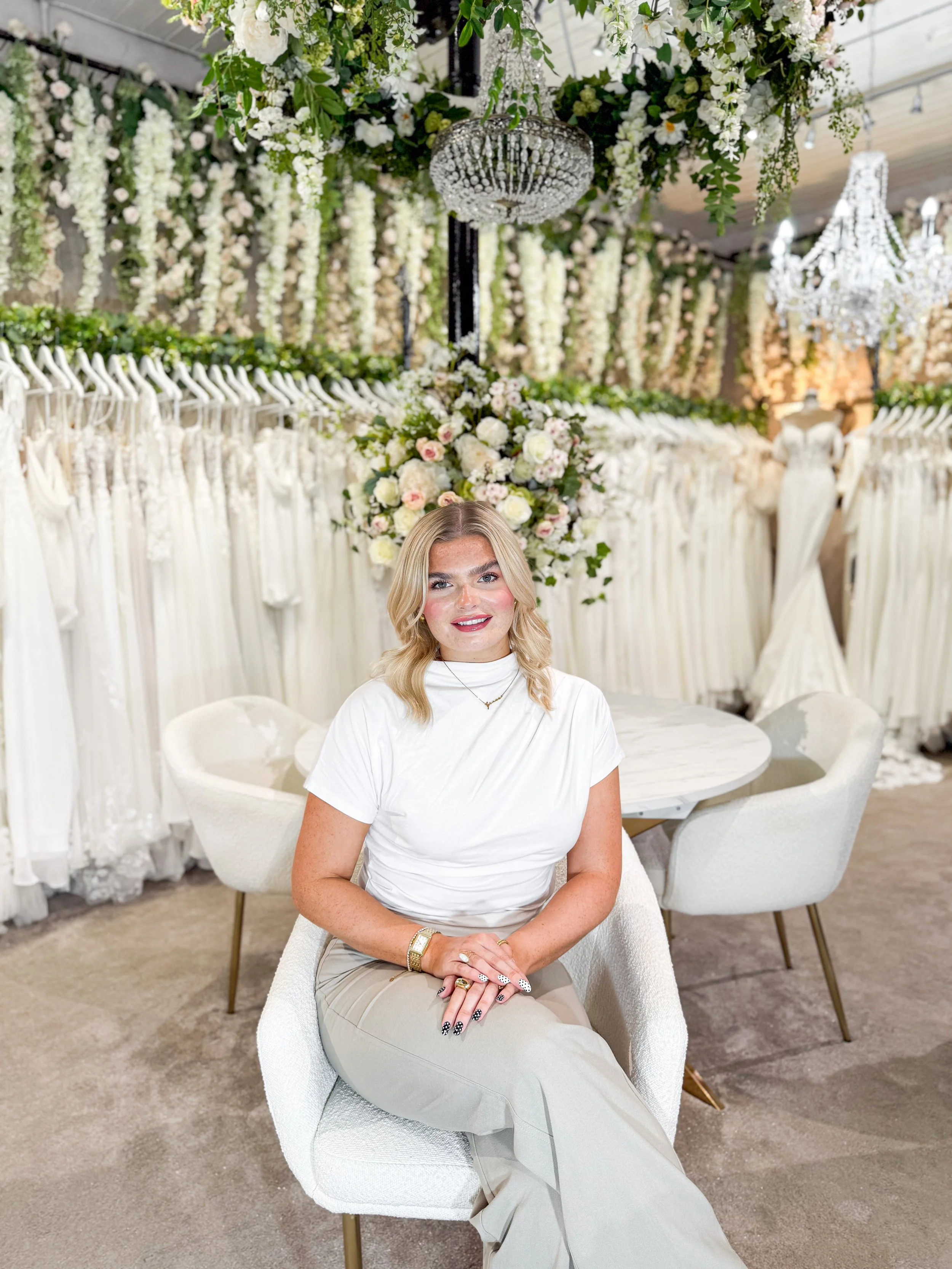 A woman standing in front of wedding dresses and mannequins in a bridal boutique, with a large flower arrangement in the background.