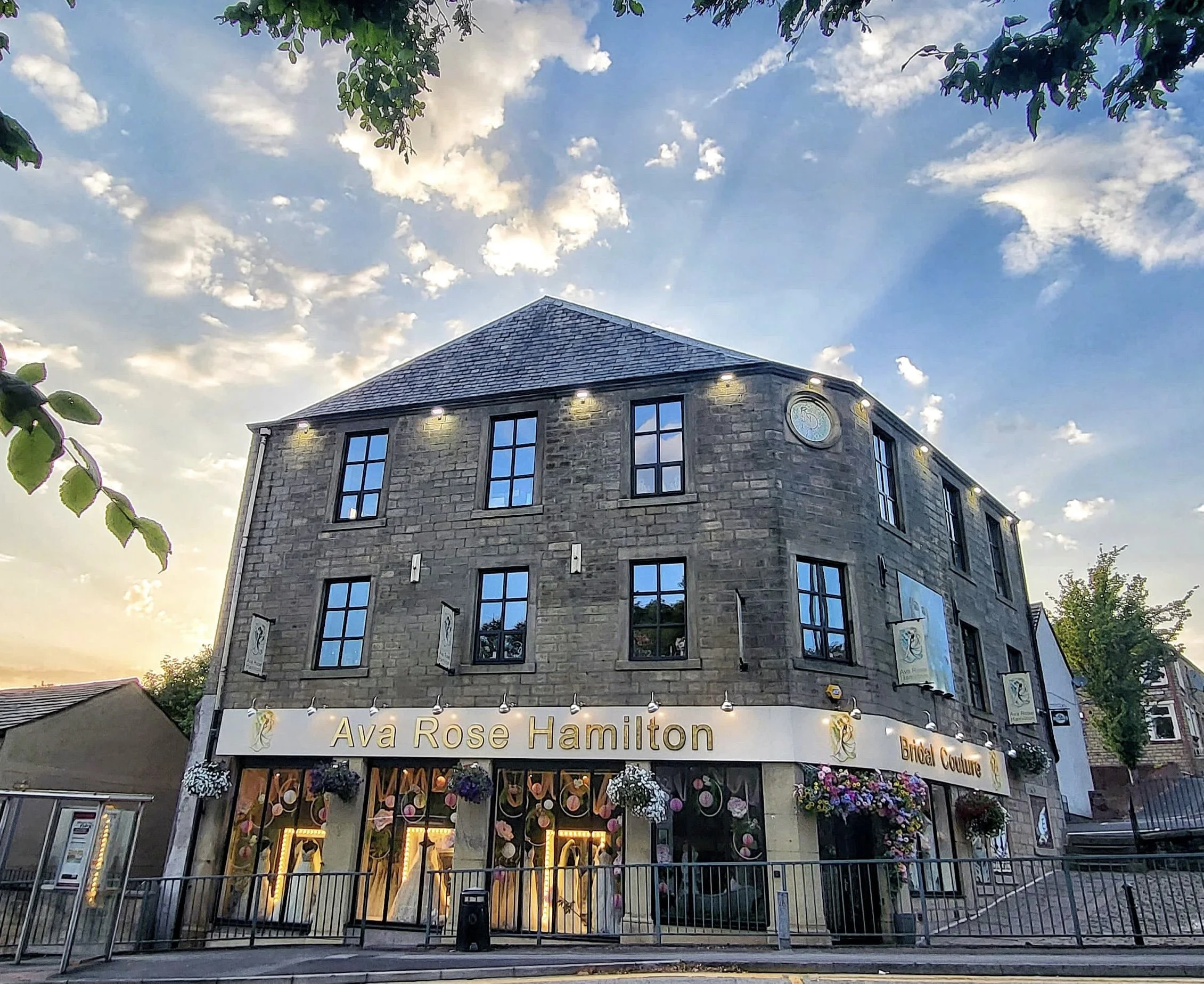 A brick building with a sign reading 'Ava Rose Hamilton Bridal Couture'. The shop window displays wedding dresses with floral decorations and hanging flower baskets outside. The building has multiple black-framed windows and some exterior lights, wit