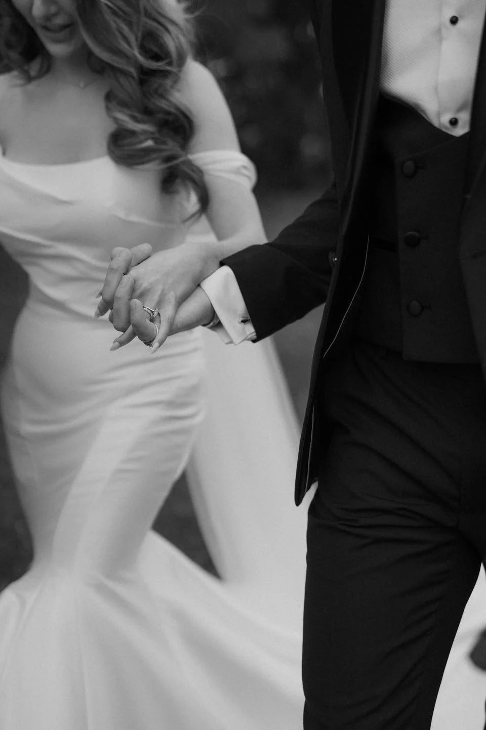 A bride and groom holding hands during their wedding dance, with the bride wearing a white off-shoulder gown and the groom in a black tuxedo.