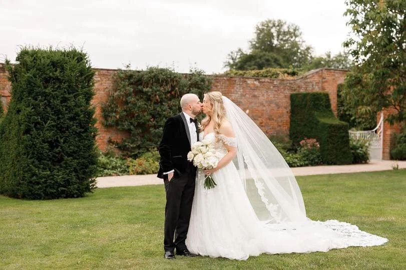 A bride and groom kissing on a lawn during their wedding, with a brick wall and greenery in the background.