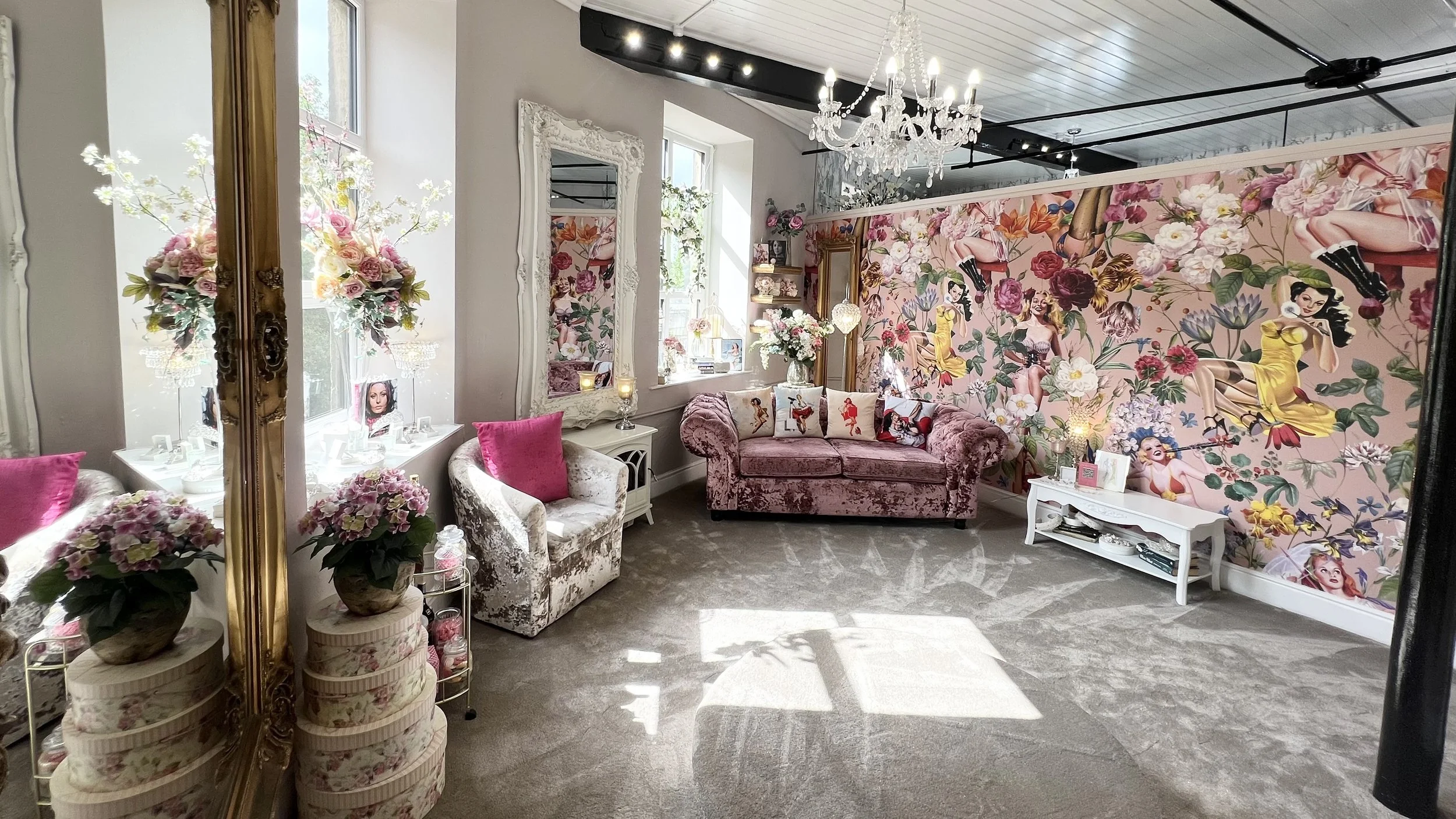 A cozy, vintage-style living room with floral wallpaper, plush pink and white velvet sofas, a chandelier, large windows with floral arrangements, and decorative pillows, creating an elegant and feminine atmosphere.