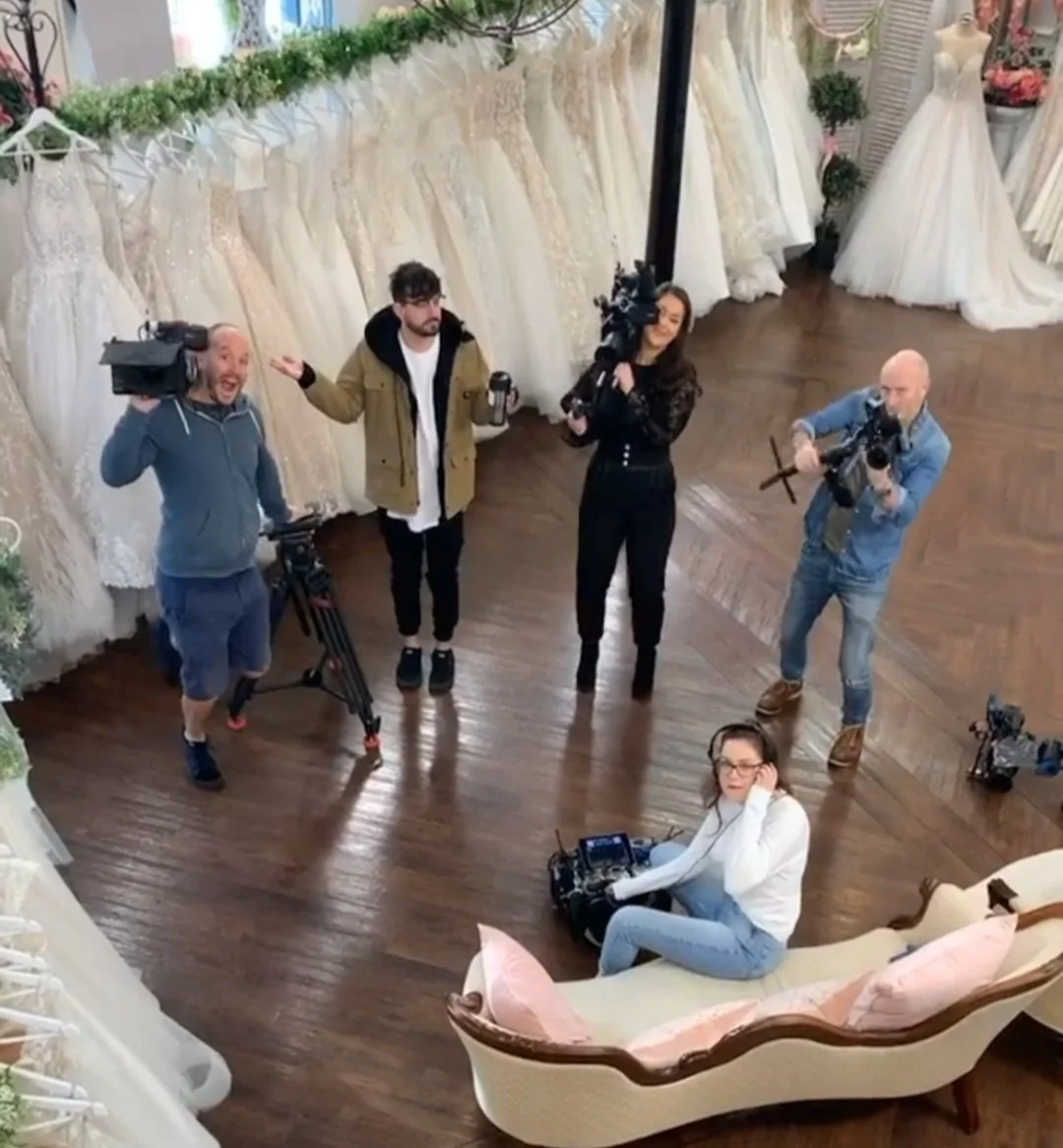 A group of five people, including three camera operators, a woman sitting on a vintage couch with a camera, and a man standing with a coffee cup, in a bridal gown shop with wedding dresses hanging on the wall.