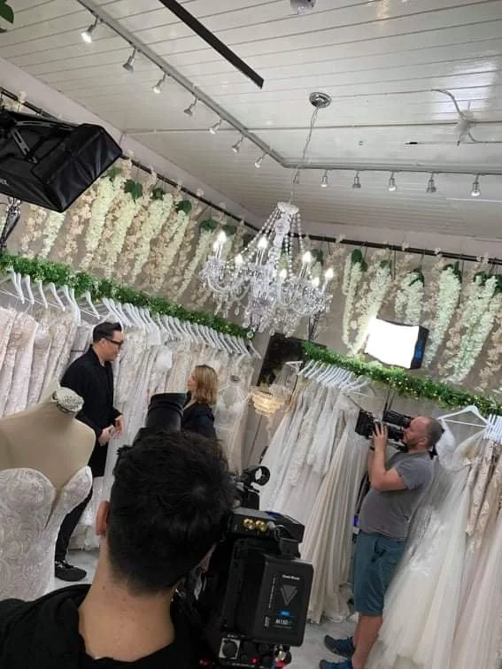 Two people are being filmed in a bridal store surrounded by wedding dresses and decorations, with a chandelier hanging from the ceiling.