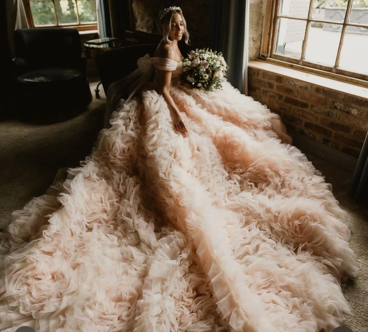 A bride sitting near a window, wearing a voluminous ruffled wedding gown and holding a bouquet of flowers.