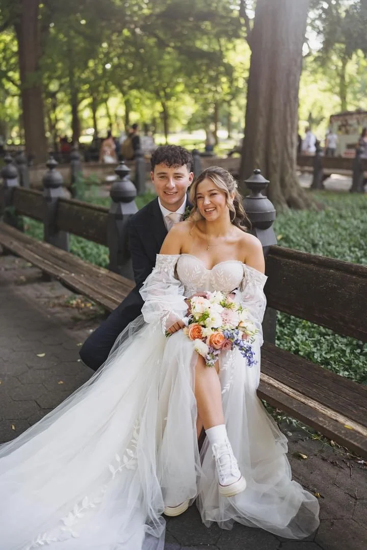 A newlywed couple sitting on a park bench surrounded by greenery, with the bride holding a bouquet of flowers and wearing a white wedding dress with off-the-shoulder sleeves, and the groom in a dark suit. They are smiling happily.