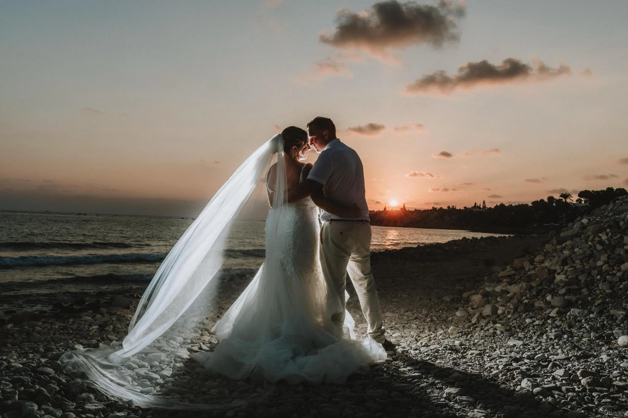 A bride and groom standing close together on a rocky beach at sunset, sharing a tender moment. The bride is wearing a white wedding dress with a long veil, and the groom is dressed in light-colored pants and a shirt. The sun is setting behind them, c
