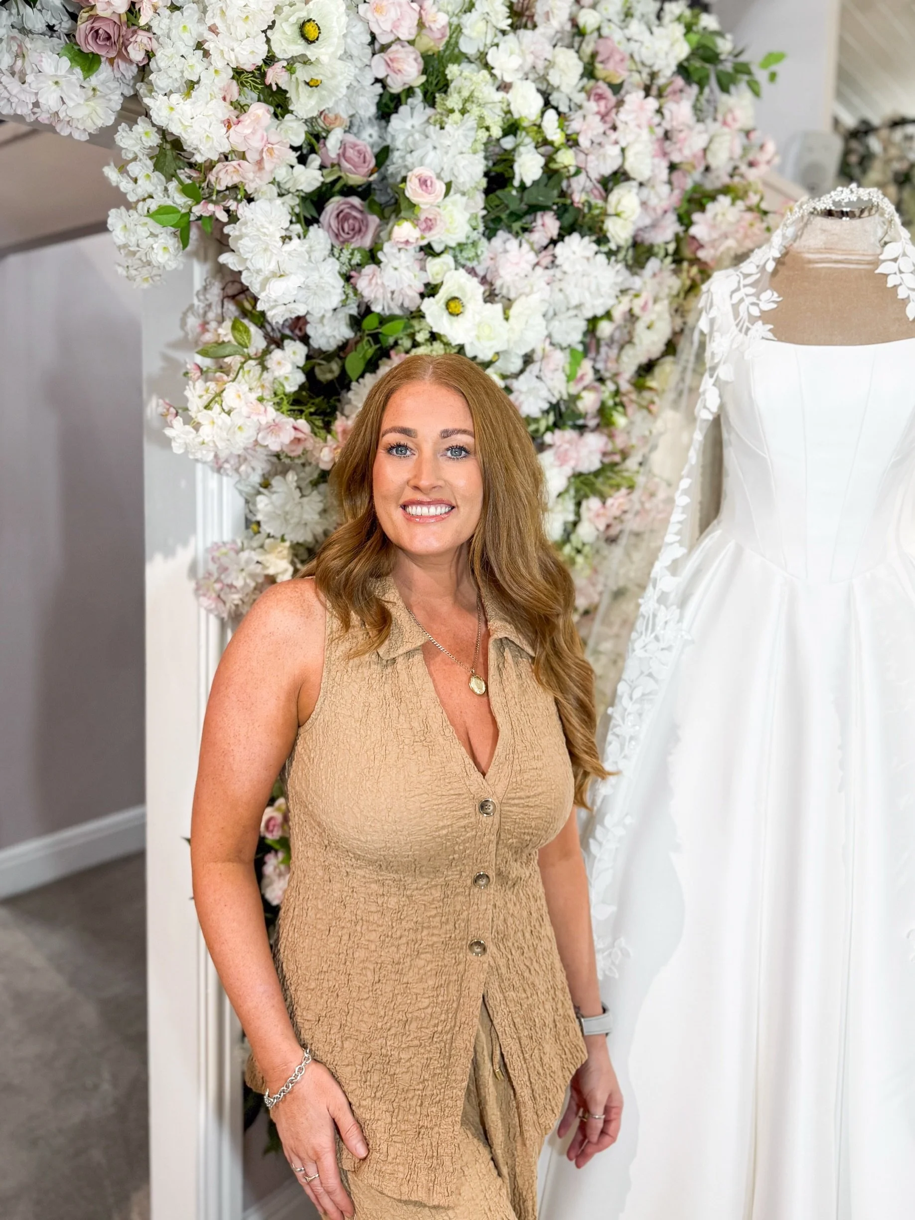 A woman standing next to a bridal dress on a mannequin, surrounded by other white wedding dresses in a boutique.