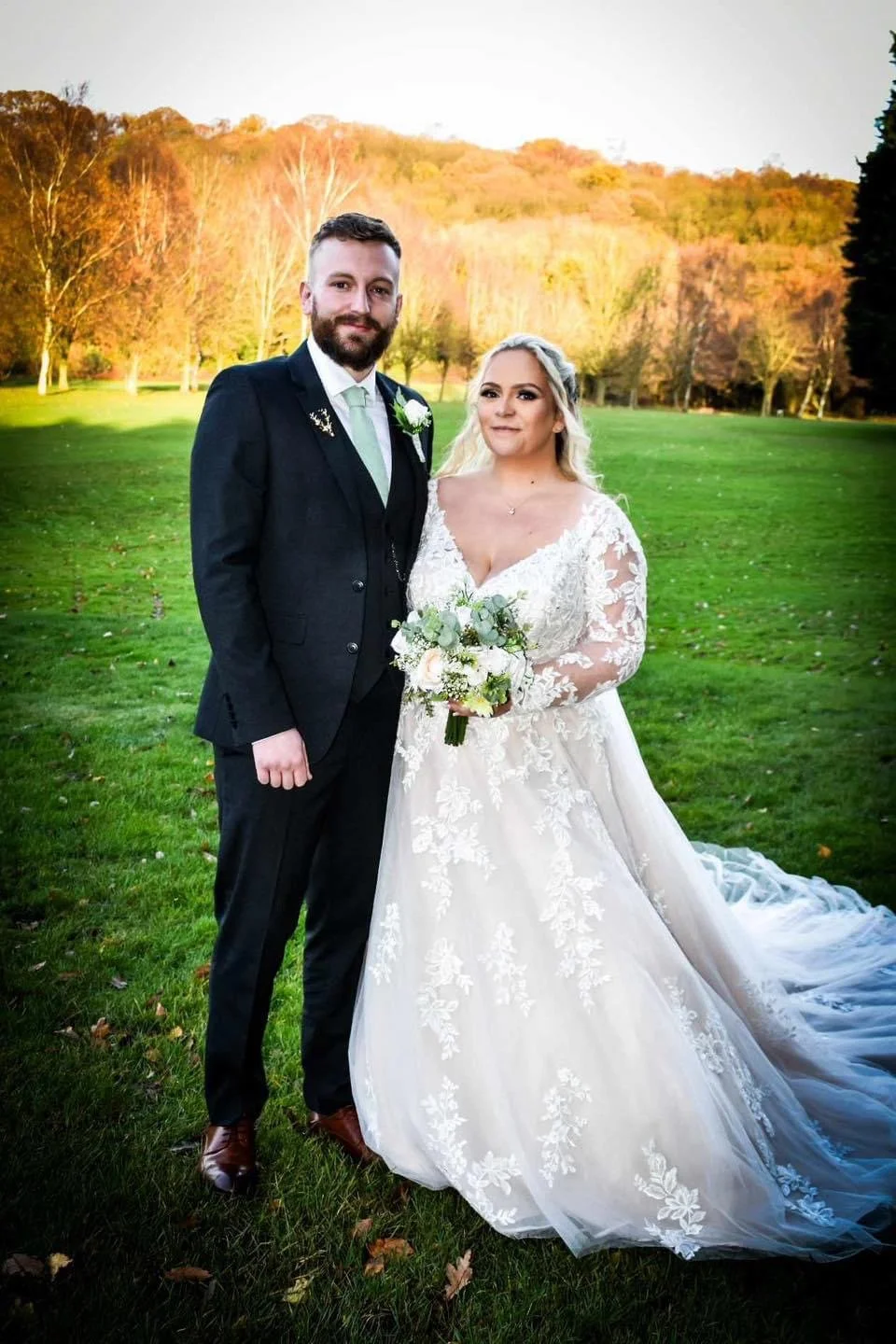A bride and groom standing outdoors in a grassy field with trees and mountains in the background during sunset, dressed in wedding attire, with the bride holding a bouquet of flowers.