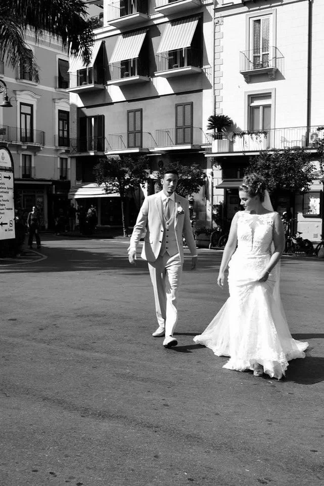 A black and white photo of a bride in a wedding gown and a groom in a suit walking on a city street.