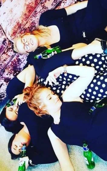 Three women lying on the floor in a casual setting, surrounded by green glass bottles, some holding bottles, with one woman wearing glasses and polka dot shirt, all appearing relaxed.