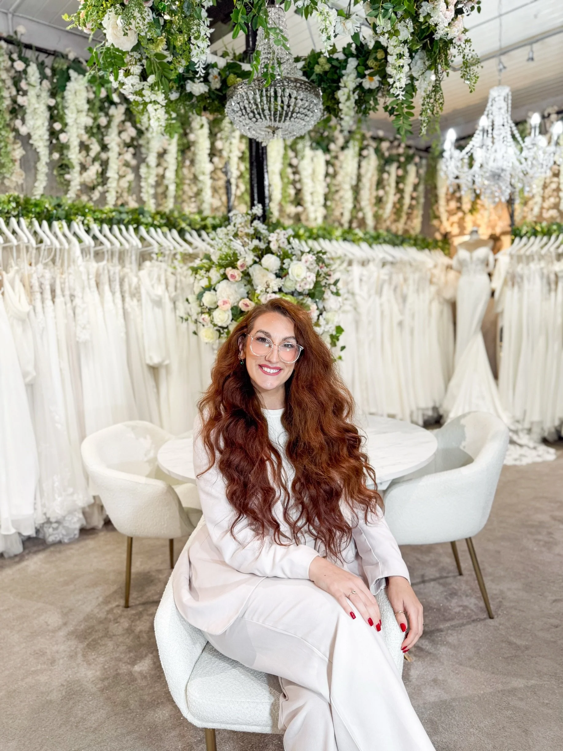 A woman with long, curly hair and glasses, dressed in a cream-colored pantsuit, standing in a room decorated for a wedding or event, next to a large floral arrangement.
