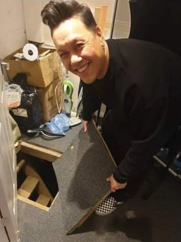Person smiling and kneeling while holding a piece of a cardboard box in a cluttered room.