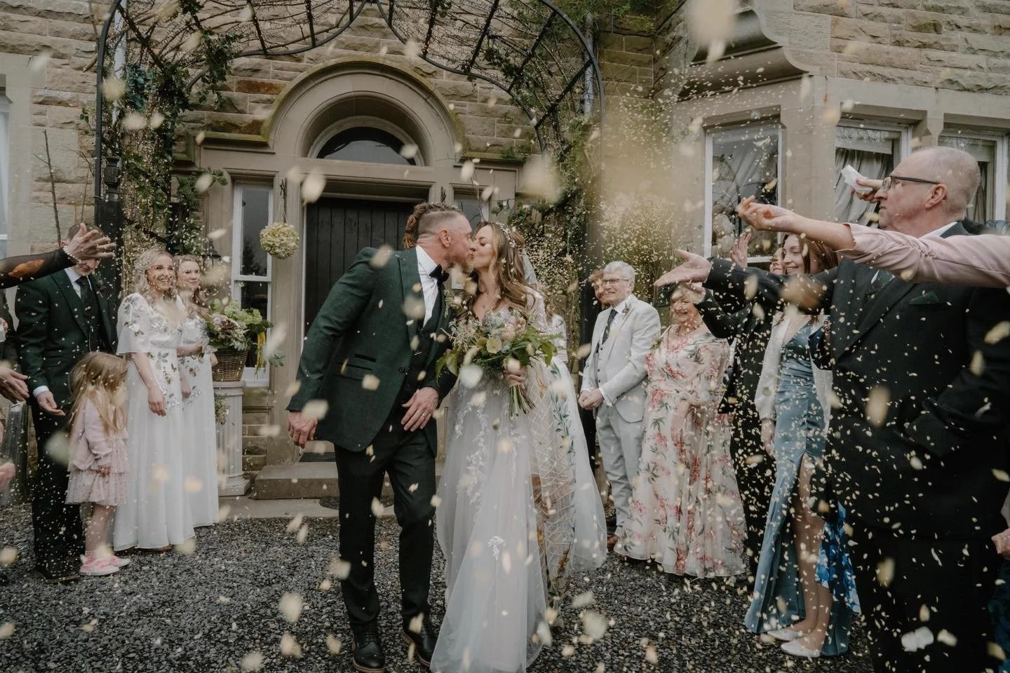 A newlywed couple kissing amid celebration, surrounded by friends and family throwing confetti outside a stone building.