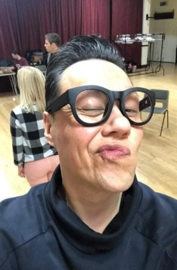 Person with short black hair, wearing glasses and a black jacket, posing with a playful expression in a room with other people and red curtains in the background.