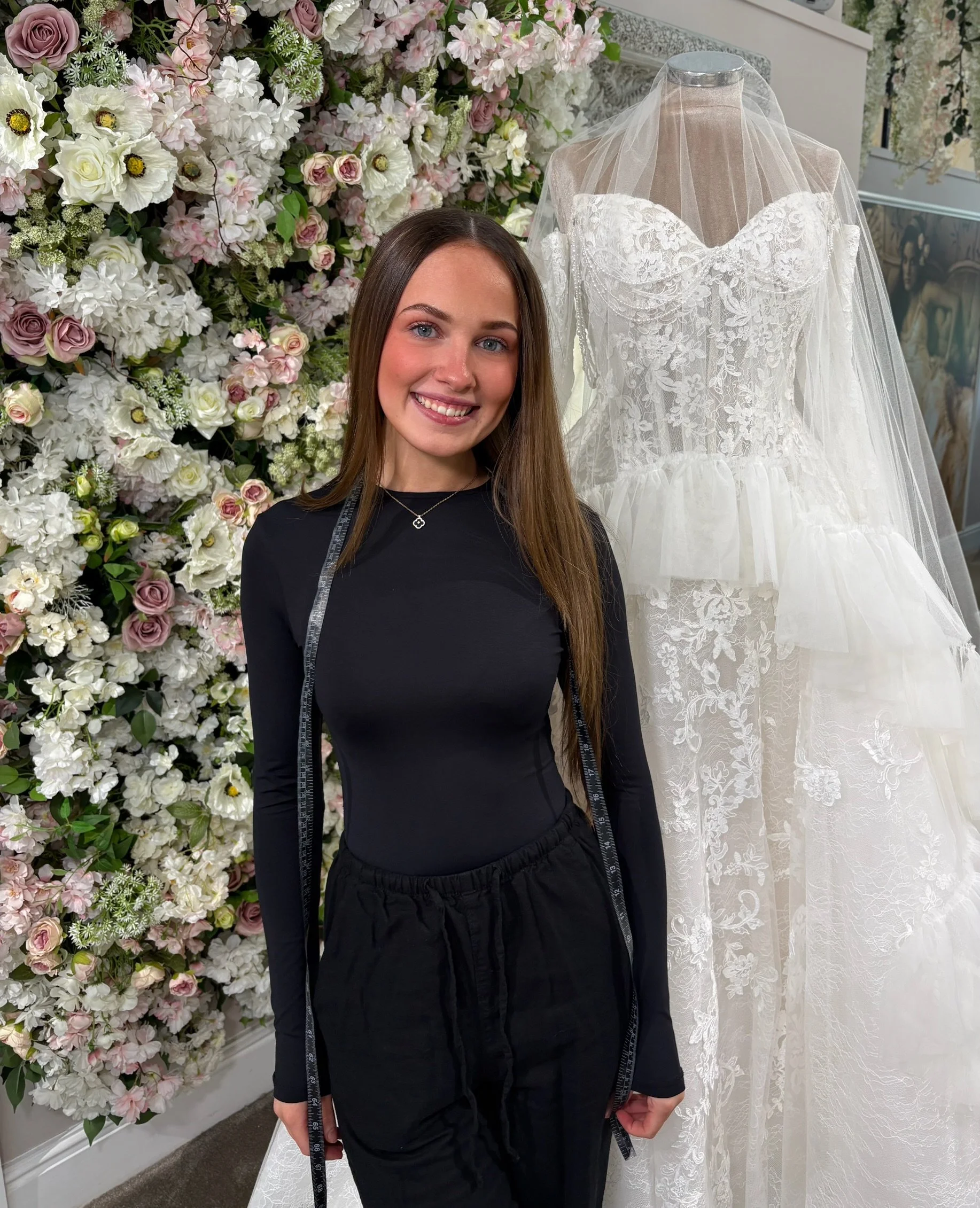 A woman with long brown hair smiling in front of wedding dresses and a floral arrangement.
