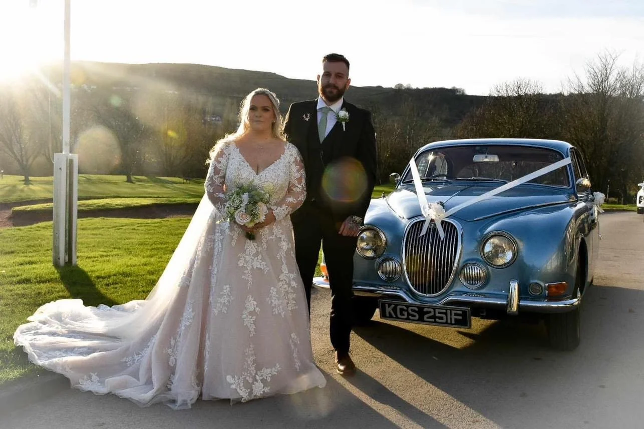 Bride and groom standing beside a vintage blue car decorated with white ribbons and flowers on a sunny day.