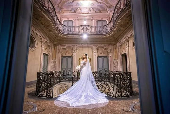 A bride in a long white wedding gown standing on a balcony in a grand, ornate hall with intricate wall moldings and a decorative ceiling, viewed through an open doorway.