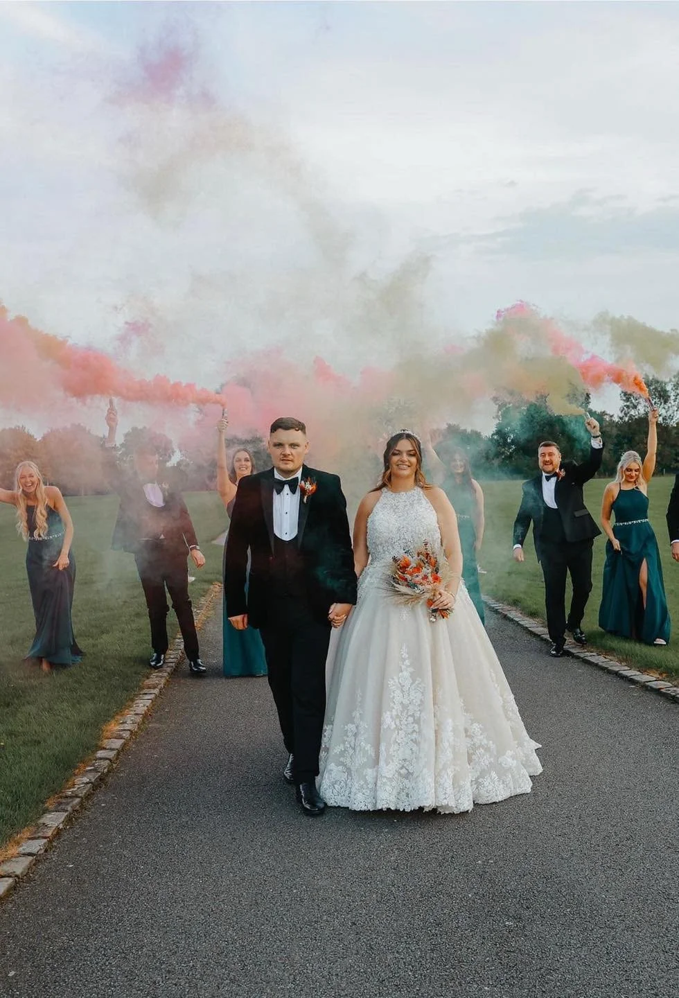 A wedding party walking along a paved path outdoors, with the bride and groom holding hands in front. The bride is wearing a white lace wedding gown and holding a bouquet, while the groom is in a black tuxedo. The bridal party behind them is holding