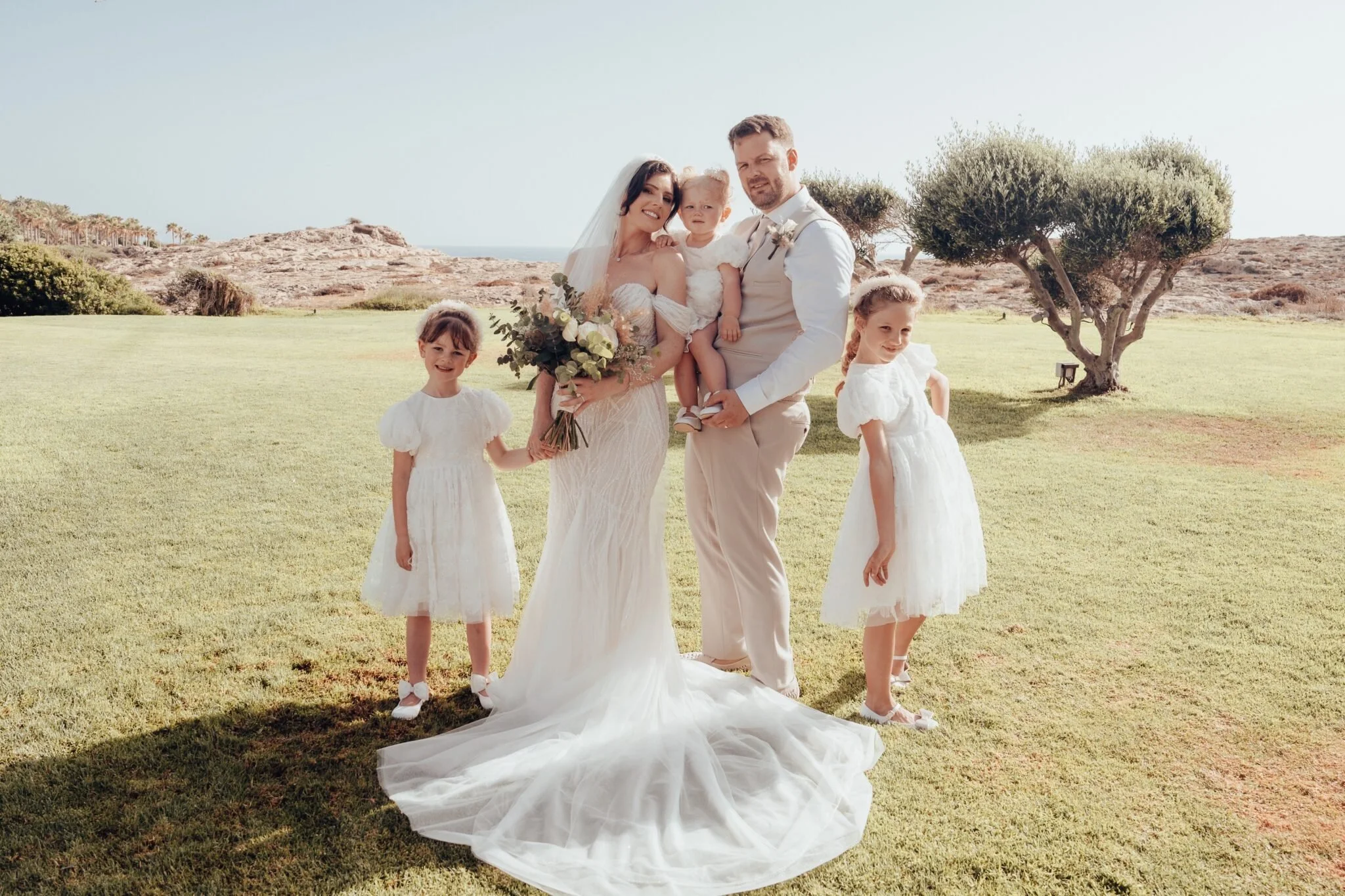 Wedding photo of a bride and groom with three young girls outdoors on a sunny day.