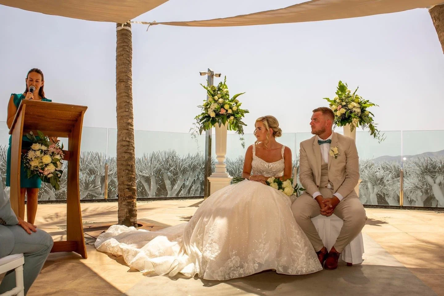 A bride in a white wedding gown and a groom in a beige suit sitting during a wedding ceremony in an outdoor setting with desert plants in the background. A woman is speaking at a podium decorated with flowers.